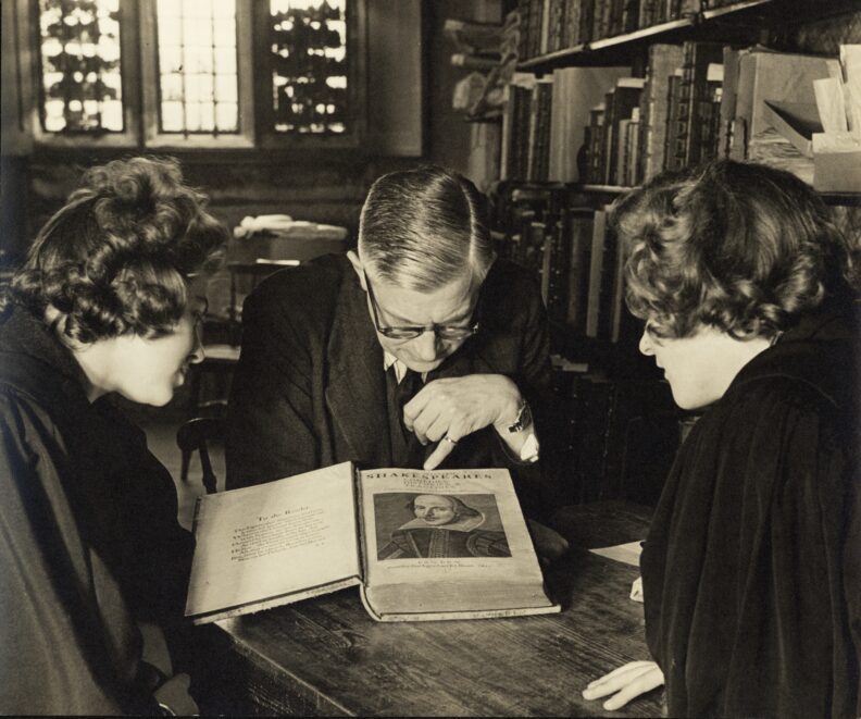 A sepia-coloured picture of a room lined with books in which a man and two women sit around a table with a copy of the First Folio in front of them. The man is pointing with a finger to the portrait on the title page.