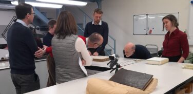 Picture showing a group of people in a conservation studio who are examining a damaged book. Another book lies closed on a cushion in the foreground. 