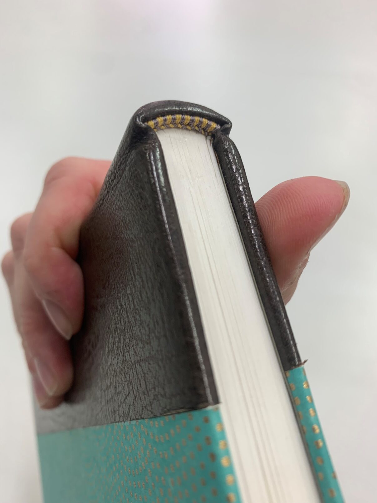 A hand holds a newly bound book for close inspection, focusing on the endband and endcap. The endband is gold and silver thread over a single core with a front bead; the end cap is lightly rounded with crisp folds. The book is bound in half leather with gold-dotted green paper.