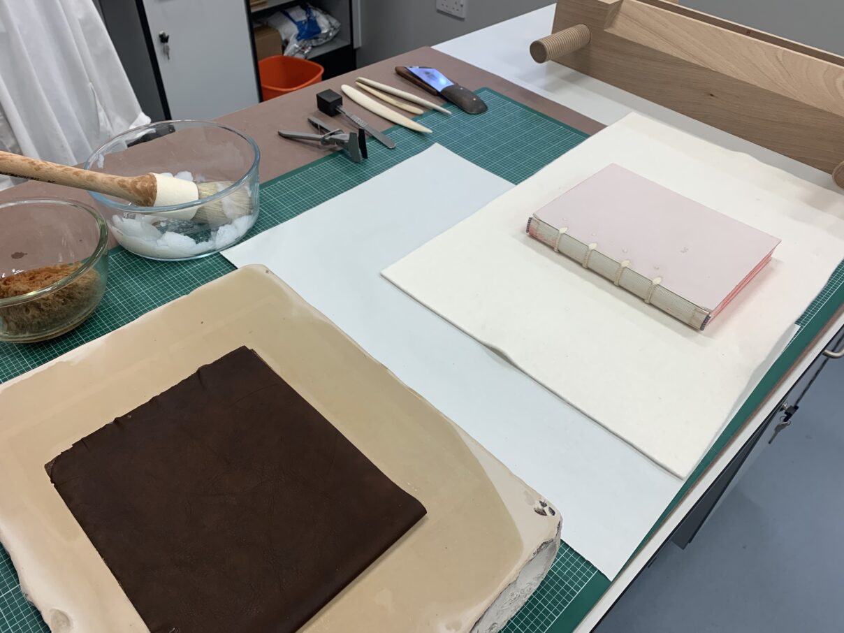 A bookbinder's bench is set up for covering. In the fore ground is a wet lithostone with pared leather. Beside it is a forwarded textblock on a felt. Behind them are a sea sponge, paste brush in a paste bowl, band nippers, a scalpel, various bone folders, and a French paring knife. In the background is a finishing press.